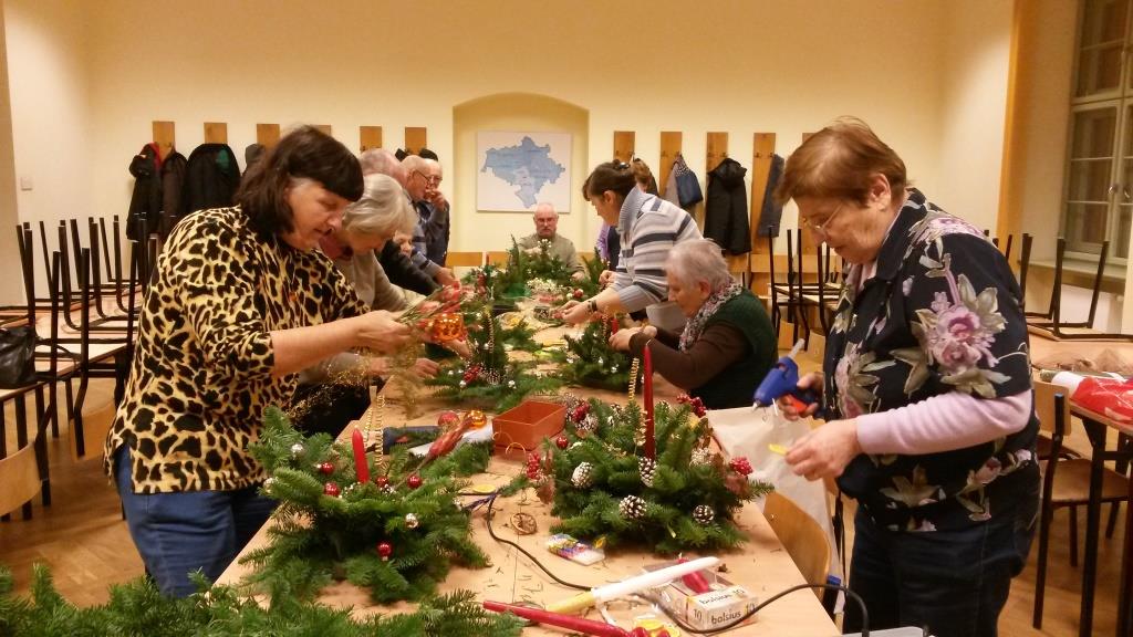 Grupa seniorów po obu stronach dużego stołu przygotowuje stroiki świąteczne