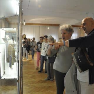 Kobieta i mężczyzna oglądają wystawę w muzeum, męzczyzna wskazuje na gablotę palcem, w tle inni zwiedzający