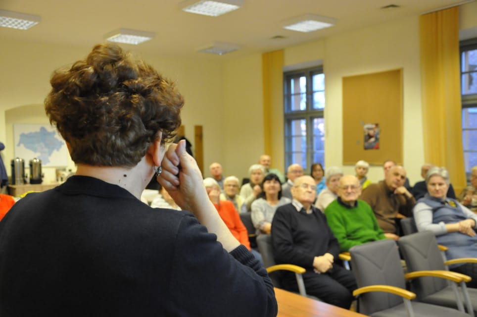 Migająca kobieta, przed nią grupa seniorów siedzi w kilku rzędach