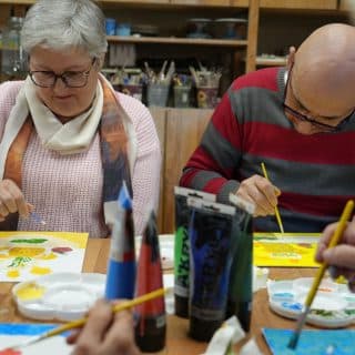 Dwoje seniorów maluje pędzlami siedząc przy stoliku w pracowni malarskiej