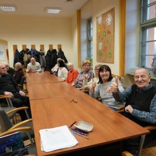 Grupa kilkunastu seniorów siedzi przy długim stole w sali