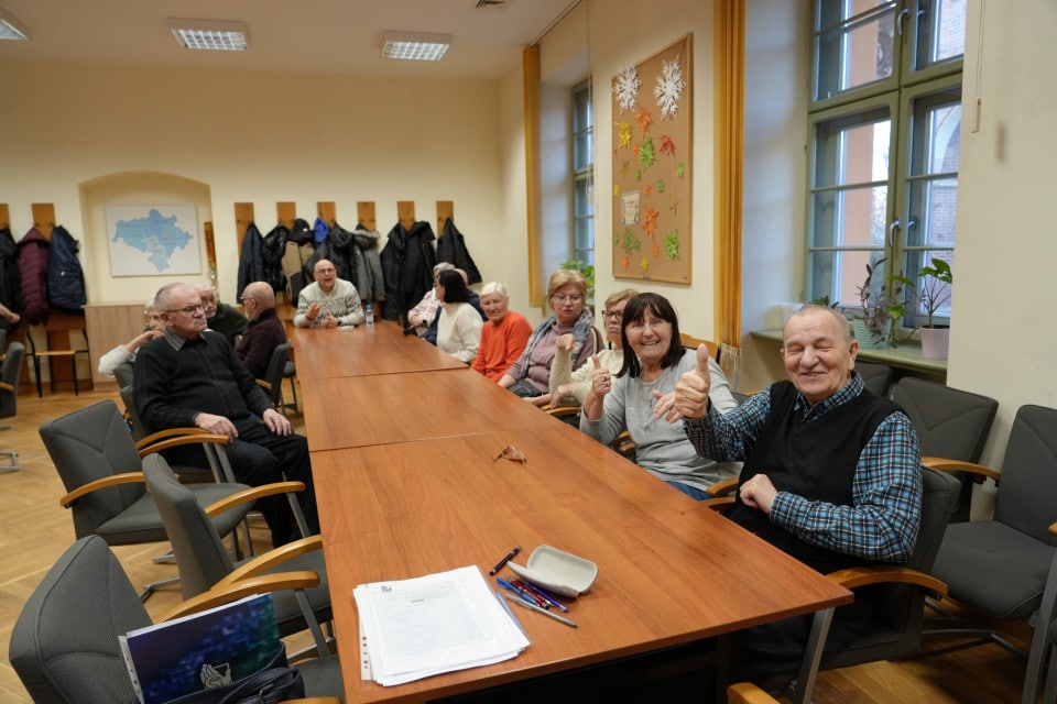 Grupa kilkunastu seniorów siedzi przy długim stole w sali