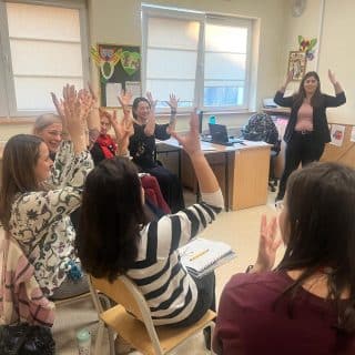 W sali kilka osób siedzi w półokręgu i macha podniesionymi rękoma