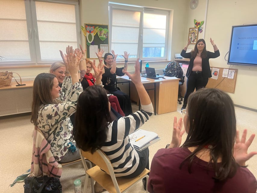 W sali kilka osób siedzi w półokręgu i macha podniesionymi rękoma