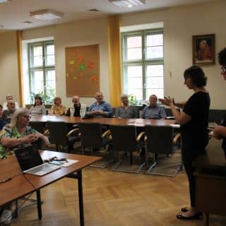 W sali siedzący seniorzy patrzą w kierunku dwóch kobiet stojących na środku