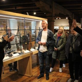 Grupa seniorów patrzy na migającego mężczyznę stojącego przy gablocie z bronią maszynową