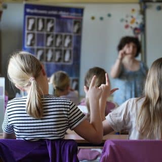 Siedzące w ławkach dzieci migaj naśladując kobietę stojącą przy tablicy z alfabetem języka migowego