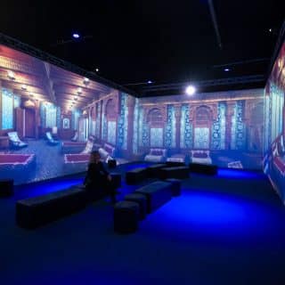 Wokół siedzącej na pufie kobiety trzy wielkie tablice, na których wyświetlone zdjęcia bogato wyposażonego pokoju