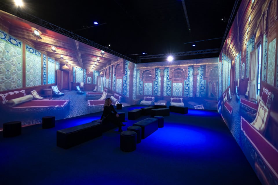 Wokół siedzącej na pufie kobiety trzy wielkie tablice, na których wyświetlone zdjęcia bogato wyposażonego pokoju