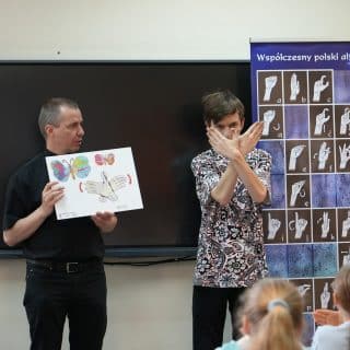 W sali szkolnej dzieci patrzą na dwóch mężczyzn - pierwszy trzyma planszę, drugi miga