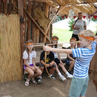 Chłopiec strzela z łuku pod opieką kobiety, w tle grupa dzieci