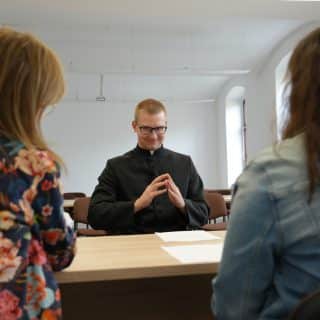 Migający młody kapłan przy stole naprzeciw dwóch kobiet