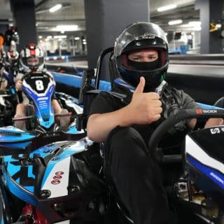 Kierowca gokarta z kciukiem podniesionym do góry, w tle inne gokarty