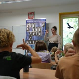 Grupa dzieci miga w sali lekcyjnej naśladując stojącą przed nimi migającą kobietę