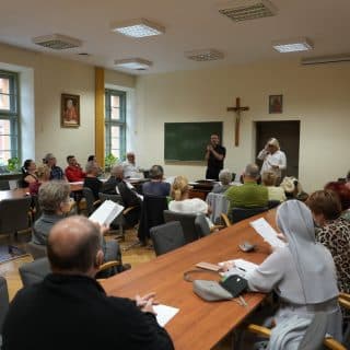 Zdjęcie kilkudziesięciu osób siedzących w sali, przed którymi na środku stoją dwie osoby