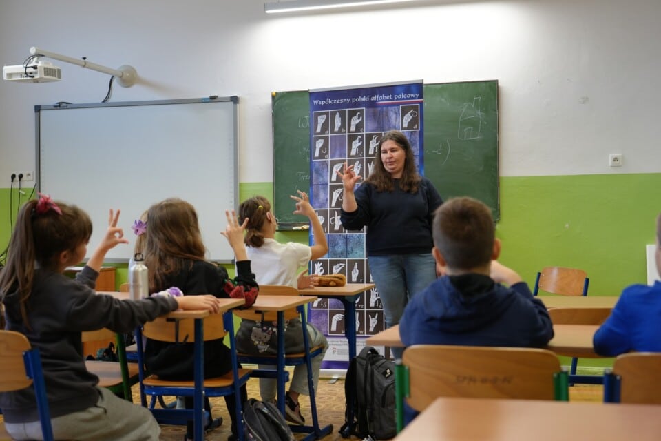 Dzieci w szkolnych ławkach naśladują migającą kobietę przy tablicy