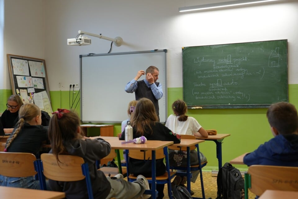 Dzieci w ławkach w sali szkolnej patrzą na mężczyznę z aparatem słuchowym