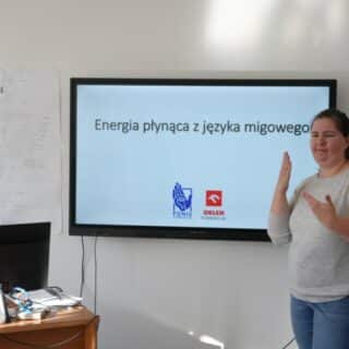 Migająca kobieta w sali przy ekranie z napisem "Energia płynąca z języka migowego"