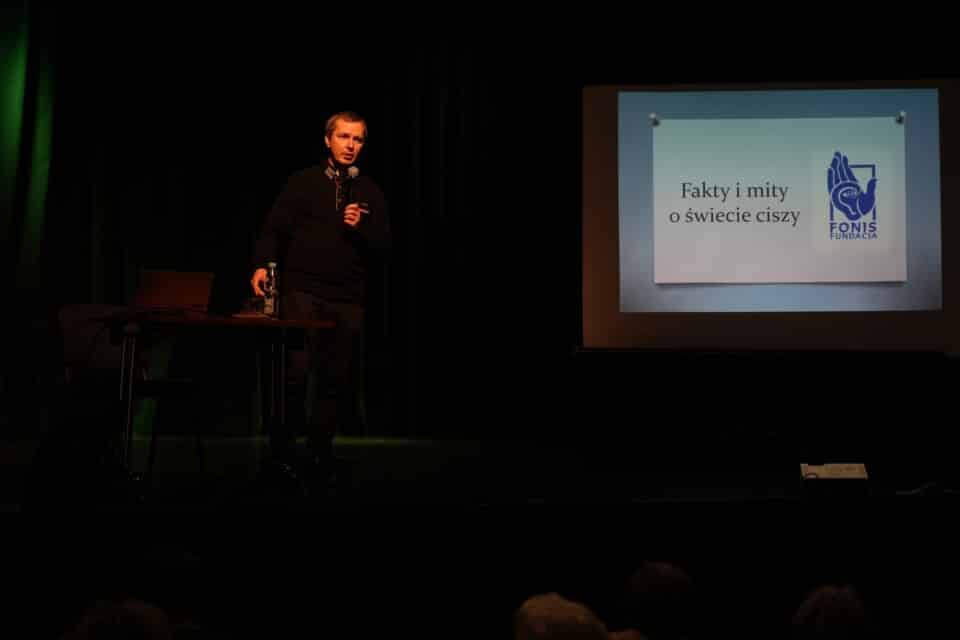 Na scenie mężczyzna przy tablicy z napisem "Fakty i mity o świecie ciszy"