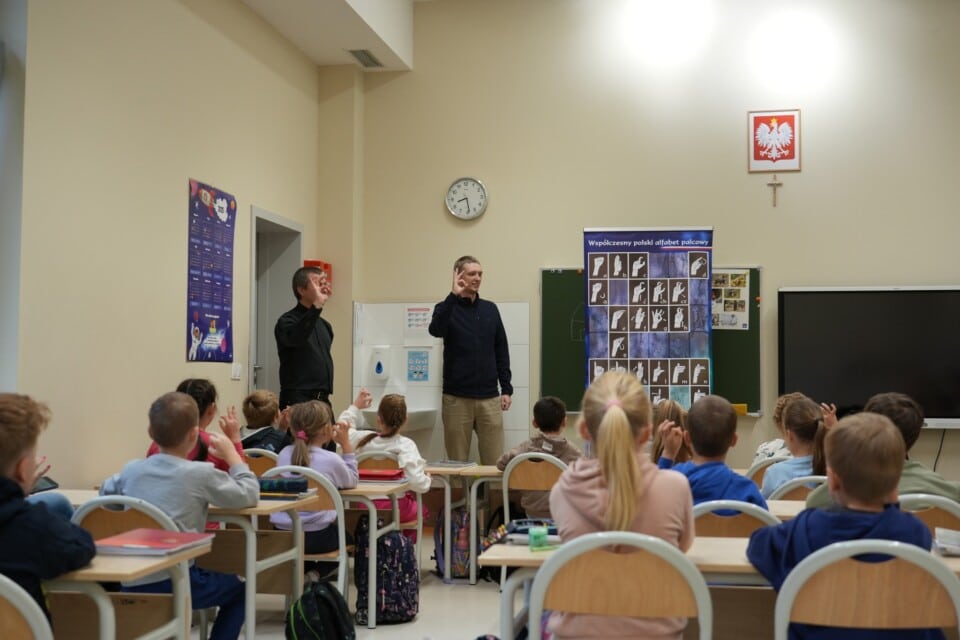 Siedzące w sali lekcyjnej dzieci naśladują dwóch stojących na środku mężczyzn