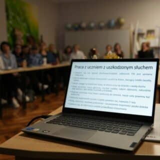 Tekst "Praca z uczniem z uszkodzonym słuchem" na laptopie. W tle w ławkach siedzą ludzie