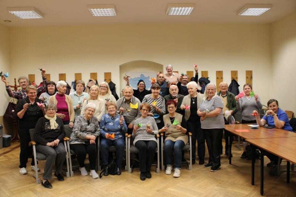 W sali zdjęcie grupowe seniorów z wykonanymi z papieru ozdobami oirgami