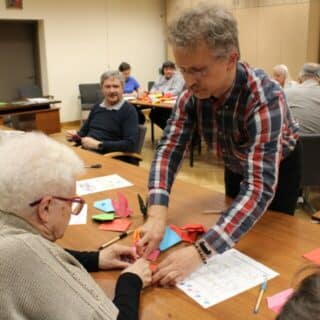 Mężczyzna pochylony nad stolikiem pomaga siedzącej kobiecie składać papier