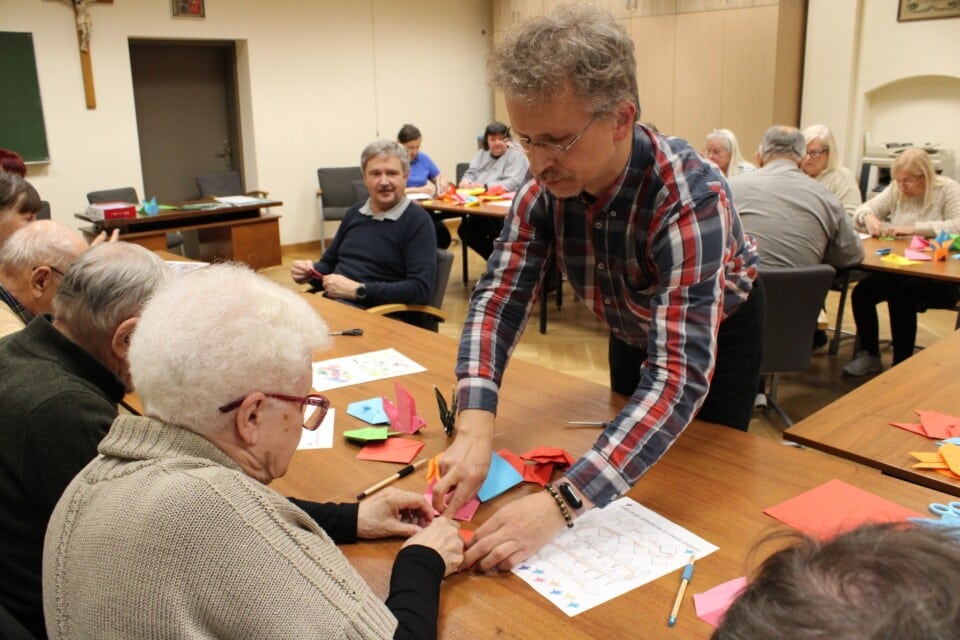 Mężczyzna pochylony nad stolikiem pomaga siedzącej kobiecie składać papier