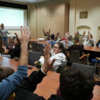 Kilkadziesiąt osób siedzących w sali podnosi ręce do góry