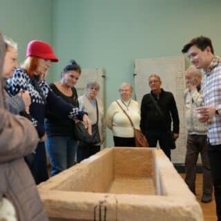 Kilka osób w pomieszczeniu stoi wokół podłużnej kamiennej rynny