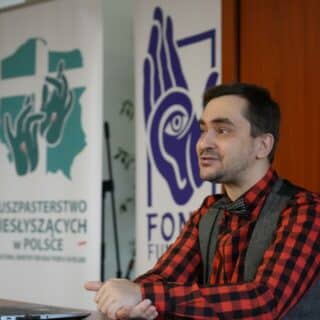 Mężczyzna przy stole na tle rollupa Duszpasterstwa Niesłyszących w Polsce i Fundacji FONIS