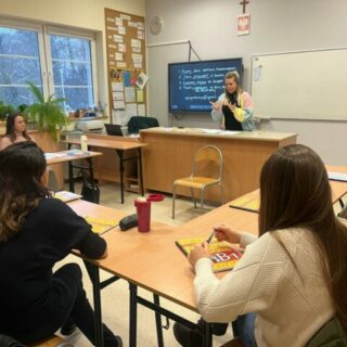 Kilka osób w sali lekcyjnej przy stolikach przed kobietą migającą przy tablicy