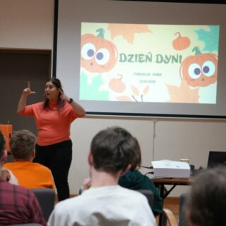 Ludzie siedzą w sali przed migającą kobietą przy ekranie z dyniową grafiką i napisem: dzień dyni